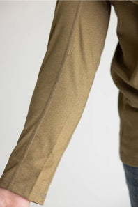 Close-up of a person's forearm wearing a textured olive-brown long-sleeve top or jacket, with a small portion of blue jeans visible at the lower right against a plain light background.