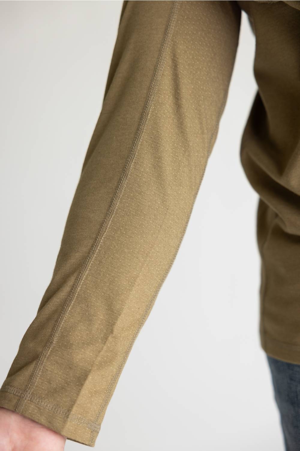 Close-up of a person's forearm wearing a textured olive-brown long-sleeve top or jacket, with a small portion of blue jeans visible at the lower right against a plain light background.