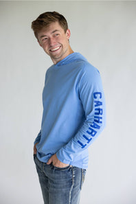 A smiling young man wearing a light blue long-sleeve hooded shirt with 'CARHART' printed vertically in dark blue on the right sleeve, paired with blue jeans, against a plain light gray background.