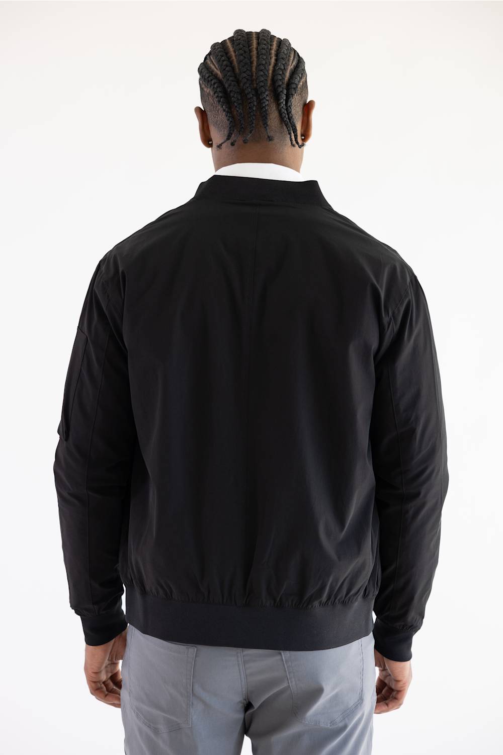 A man viewed from behind wearing a black bomber jacket, light gray trousers, and a white collared shirt, against a plain white studio background.