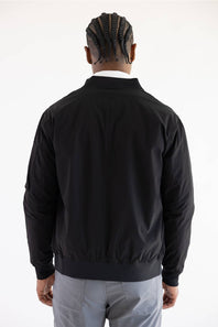 A man viewed from behind wearing a black bomber jacket, light gray trousers, and a white collared shirt, against a plain white studio background.