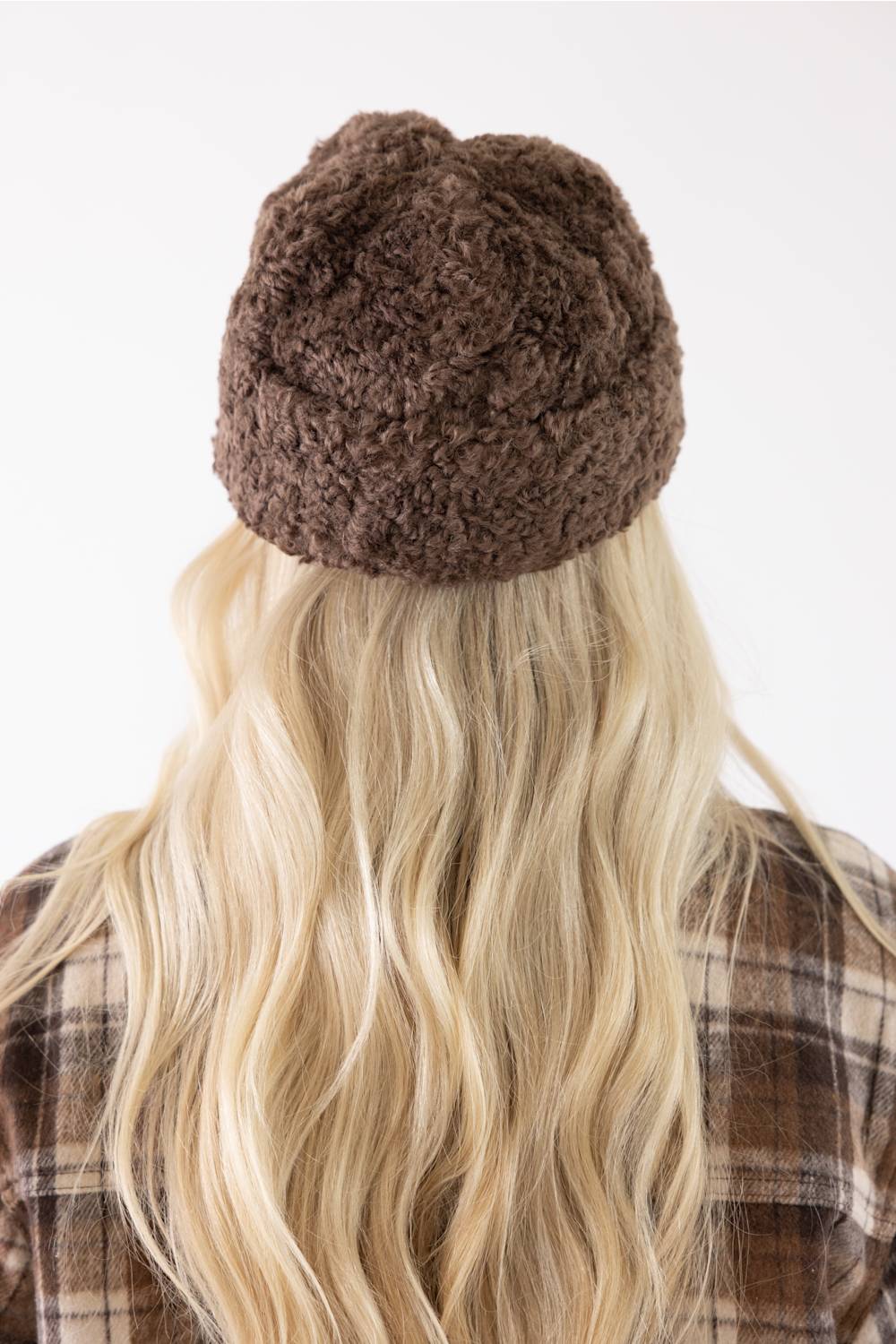 A person viewed from the back wearing a brown textured knit beanie, with long blonde hair and a brown plaid shirt against a plain white background.
