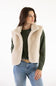 A woman modeling a cream-colored fuzzy shearling-style sleeveless vest over a dark green knit sweater and blue jeans, photographed against a plain white studio background.