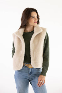 A woman modeling a cream-colored fuzzy shearling-style sleeveless vest over a dark green knit sweater and blue jeans, photographed against a plain white studio background.
