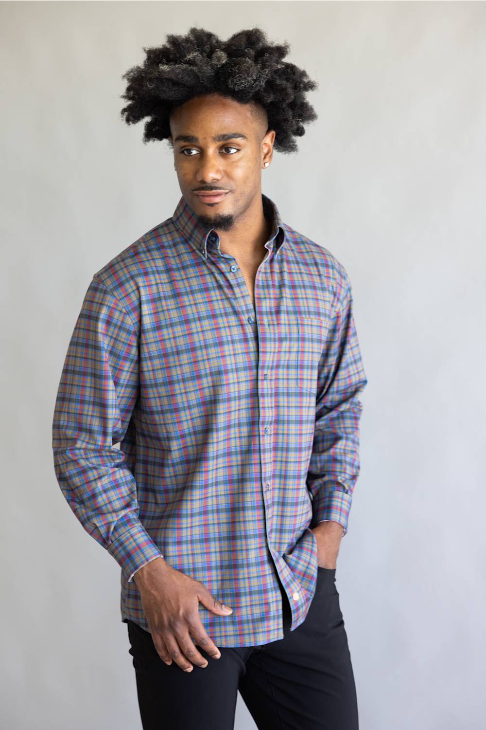 A man with an afro hairstyle wearing a long-sleeve button-down plaid shirt in blue, red, and white, paired with black pants, standing against a plain light gray background.