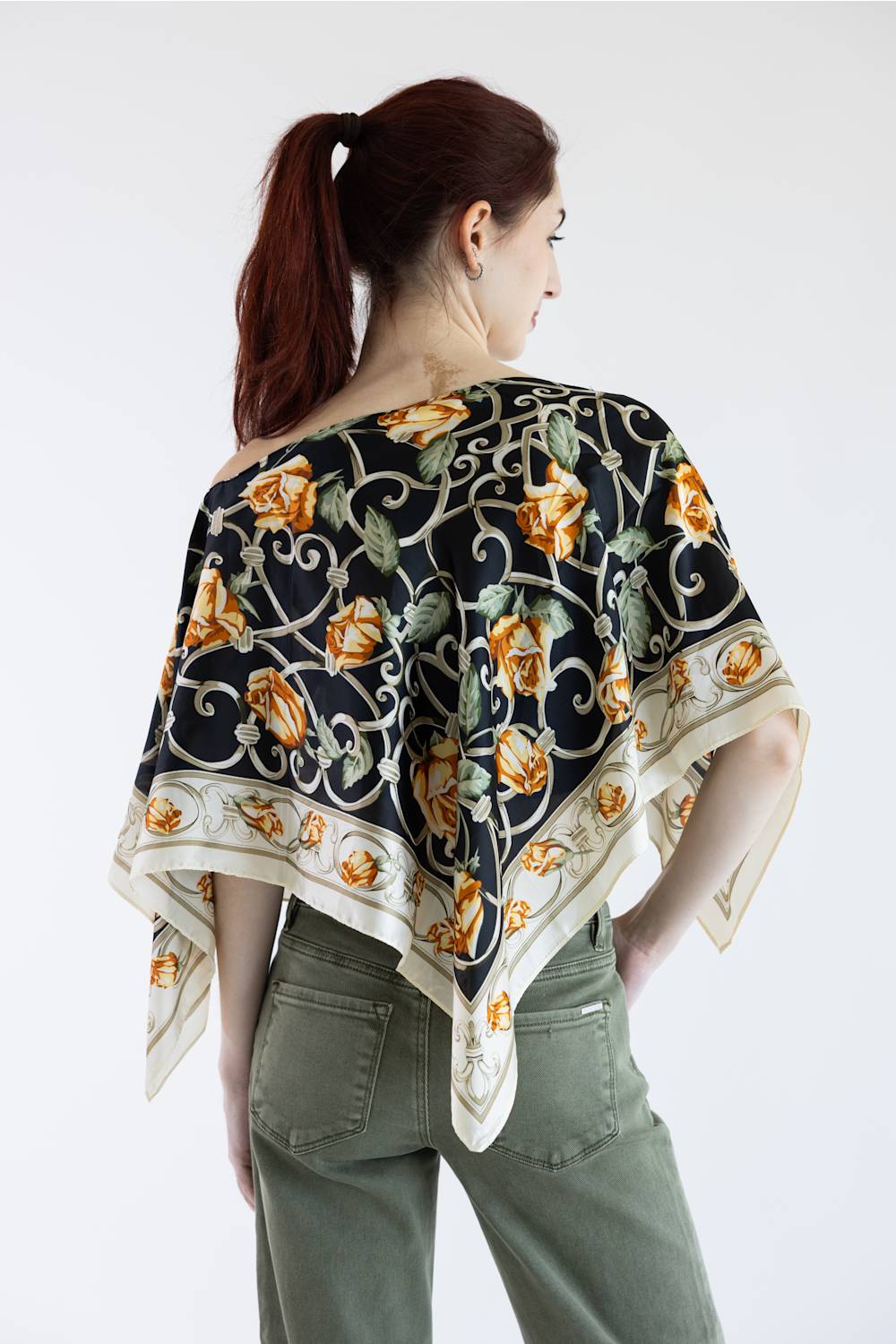 A woman shown from the back wearing a black silk scarf with a gold and green floral pattern, paired with olive green pants, against a plain white background.
