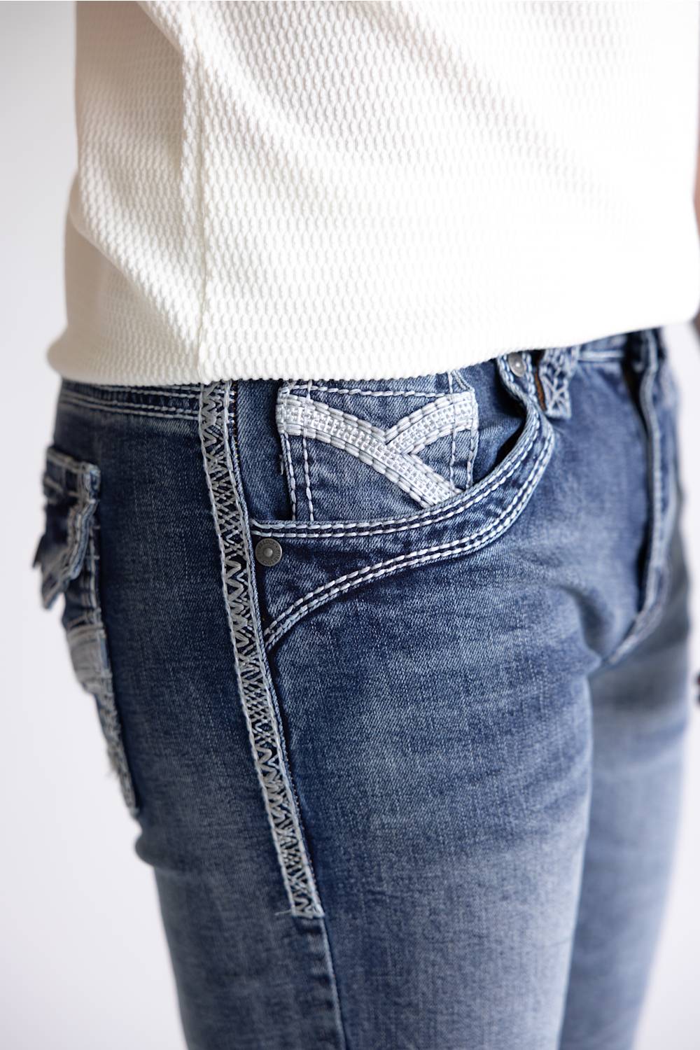 Close-up of a person wearing blue denim jeans with decorative contrast stitching and a white textured top, cropped from the waist down against a light neutral background.