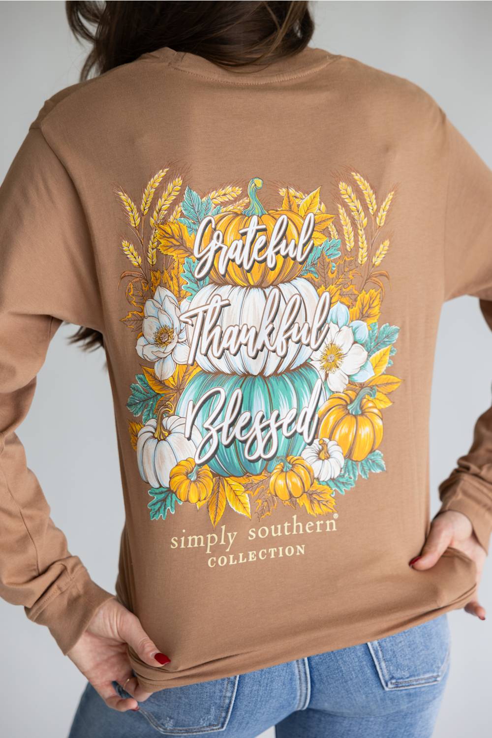 A person wearing a tan long-sleeve shirt from the back, featuring a large autumn-themed graphic with pumpkins, flowers, and the words 'Grateful Thankful Blessed' and 'simply southern COLLECTION'.