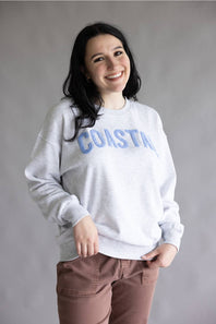 A woman smiling and wearing a light gray crewneck sweatshirt with 'COASTAL' printed across the chest in blue, paired with brown pants, against a plain gray studio background.