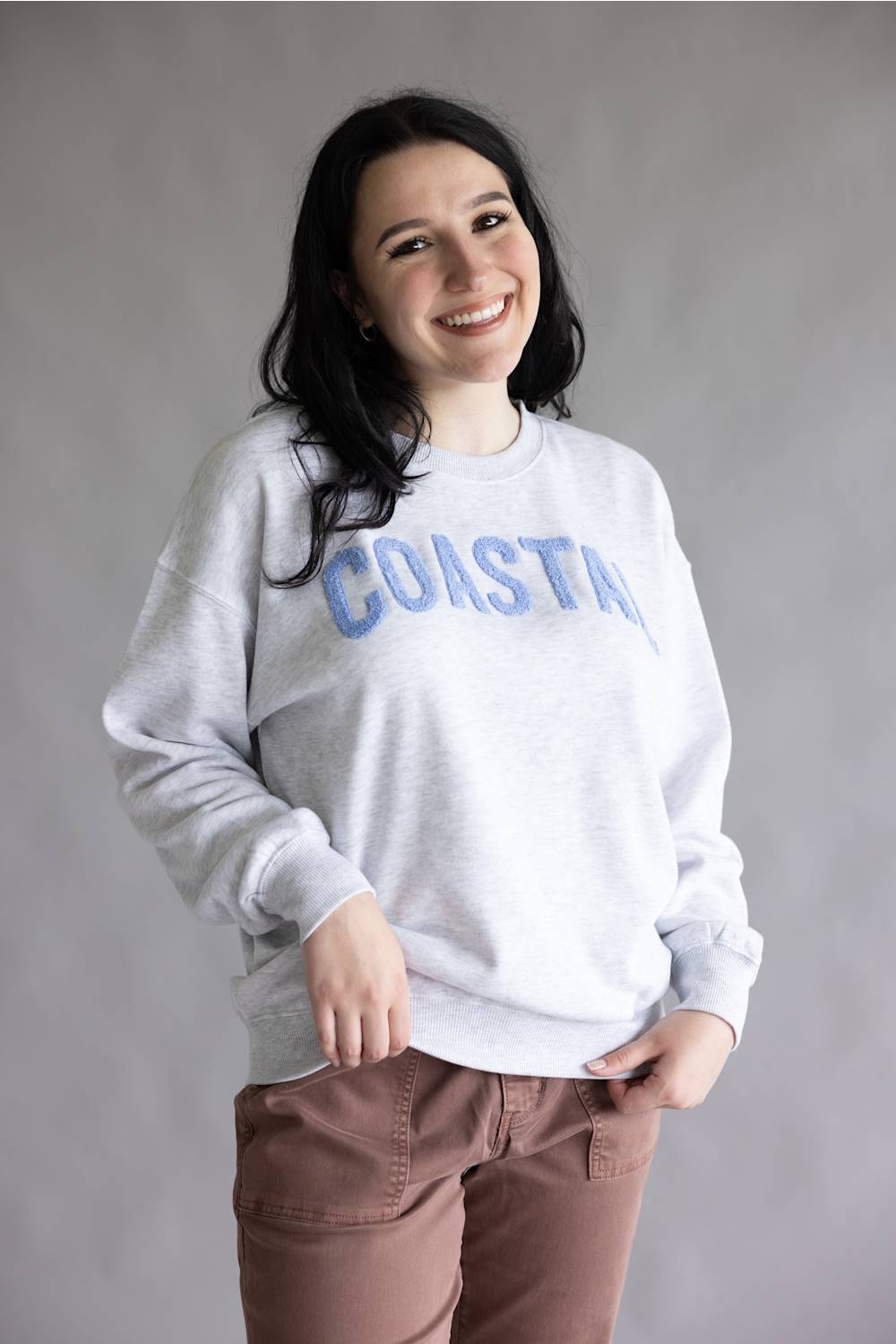 A woman smiling and wearing a light gray crewneck sweatshirt with 'COASTAL' printed across the chest in blue, paired with brown pants, against a plain gray studio background.