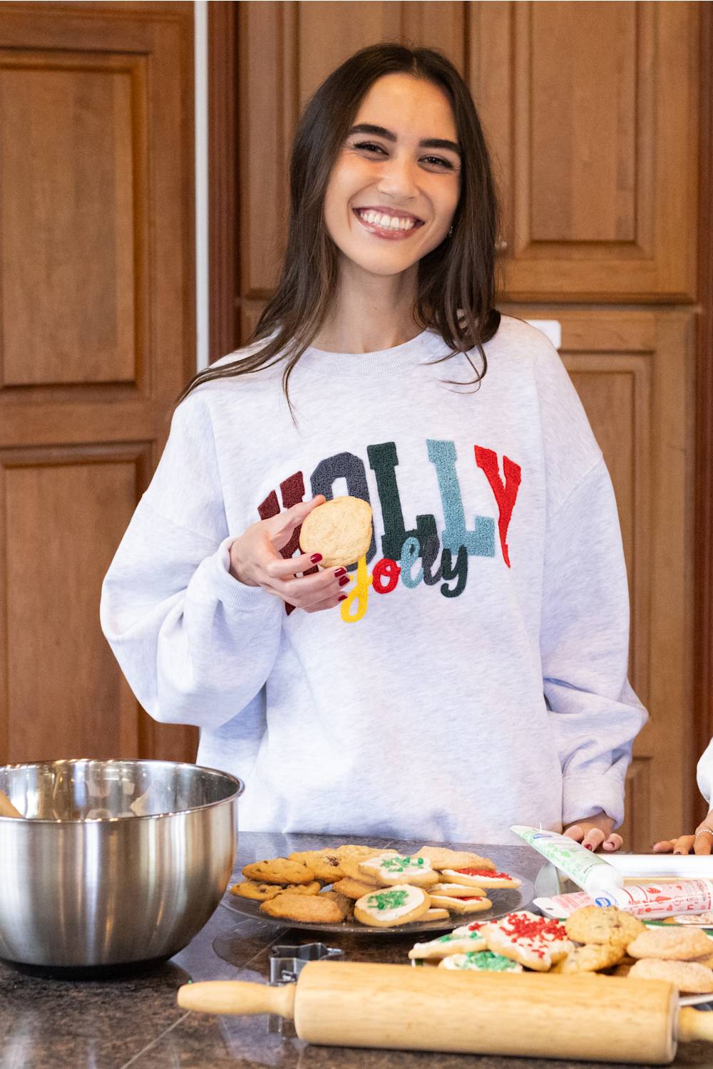 A smiling woman stands in a kitchen wearing a light gray sweatshirt with a colorful 'Holly jolly' graphic. She is holding a cookie in front of a counter with decorated cookies, a metal mixing bowl, and a wooden rolling pin.