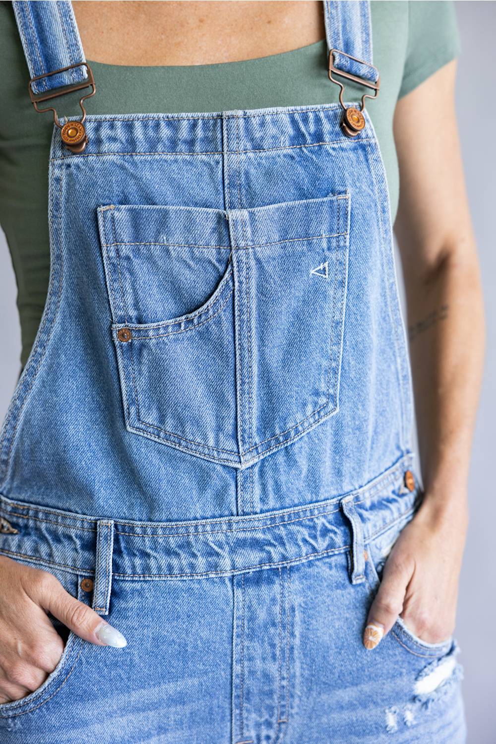Close-up of a person wearing light blue denim overall with front patch pocket, brown metal rivets, and distressed details, paired with a muted green top.