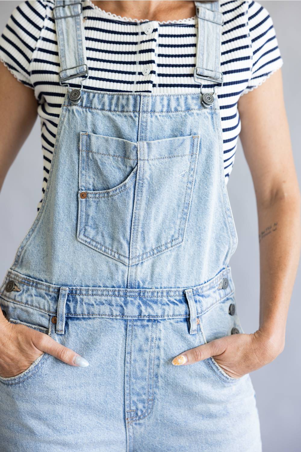 A person wearing light-wash denim overall with a black-and-white striped short-sleeve top underneath, shown from the chest down against a plain light gray background.
