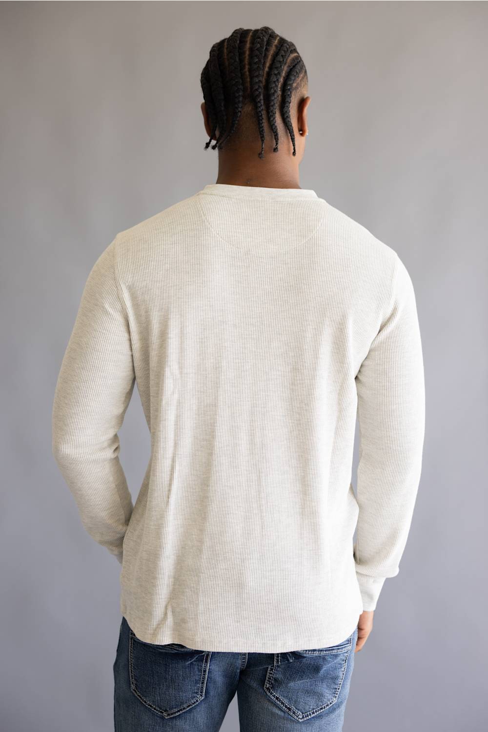 A man viewed from behind wearing a light cream-colored long-sleeve knit shirt and blue jeans, standing against a neutral gray background.