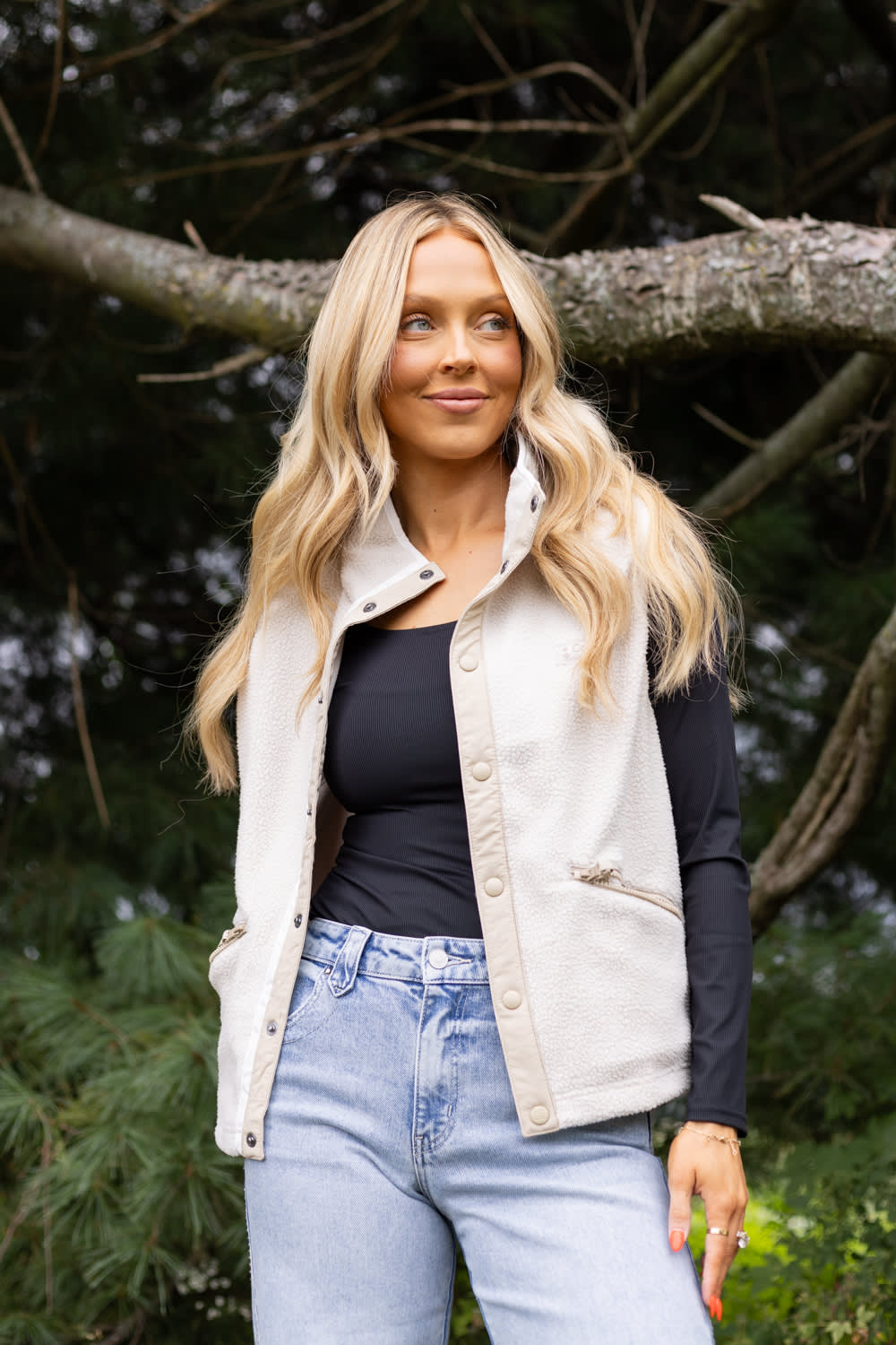 A blonde woman wearing a cream-colored sleeveless button-up vest over a black long-sleeve top and light blue jeans, standing outdoors in front of a tree branch.