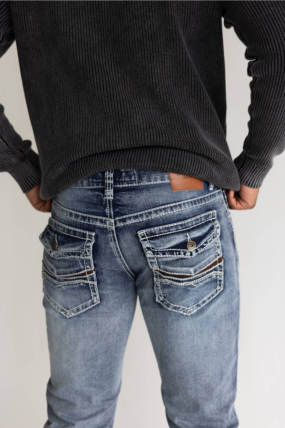 Close-up back view of a person wearing light-wash blue denim jeans with decorative stitching and a brown leather waistband, paired with a dark ribbed sweater against a plain light background.
