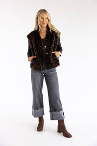 A woman modeling a brown and black leopard-print faux fur vest over a black top, paired with gray wide-leg jeans and brown ankle boots, against a plain white studio background.