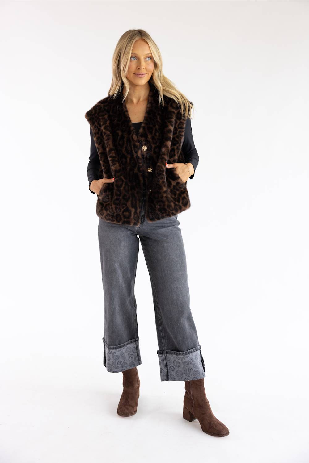 A woman modeling a brown and black leopard-print faux fur vest over a black top, paired with gray wide-leg jeans and brown ankle boots, against a plain white studio background.