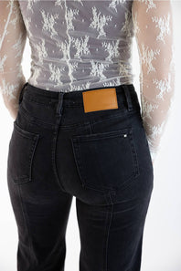 Rear view of a person wearing dark washed denim jeans with a tan leather brand patch at the back waistband, paired with a sheer white lace top against a white studio background.