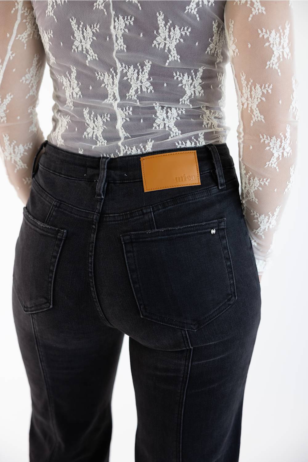 Rear view of a person wearing dark washed denim jeans with a tan leather brand patch at the back waistband, paired with a sheer white lace top against a white studio background.