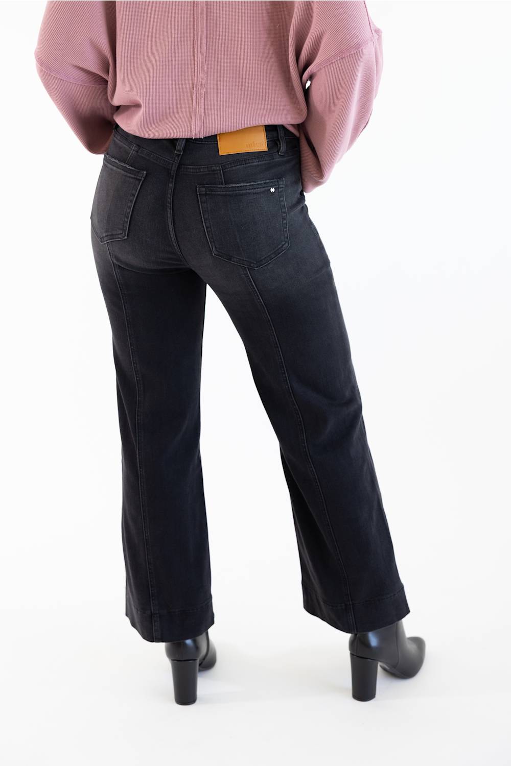 Rear view of a person wearing dark charcoal black flare jeans with a small tan leather patch at the back waistband, paired with a dusty pink top and black heeled boots, on a white studio background.