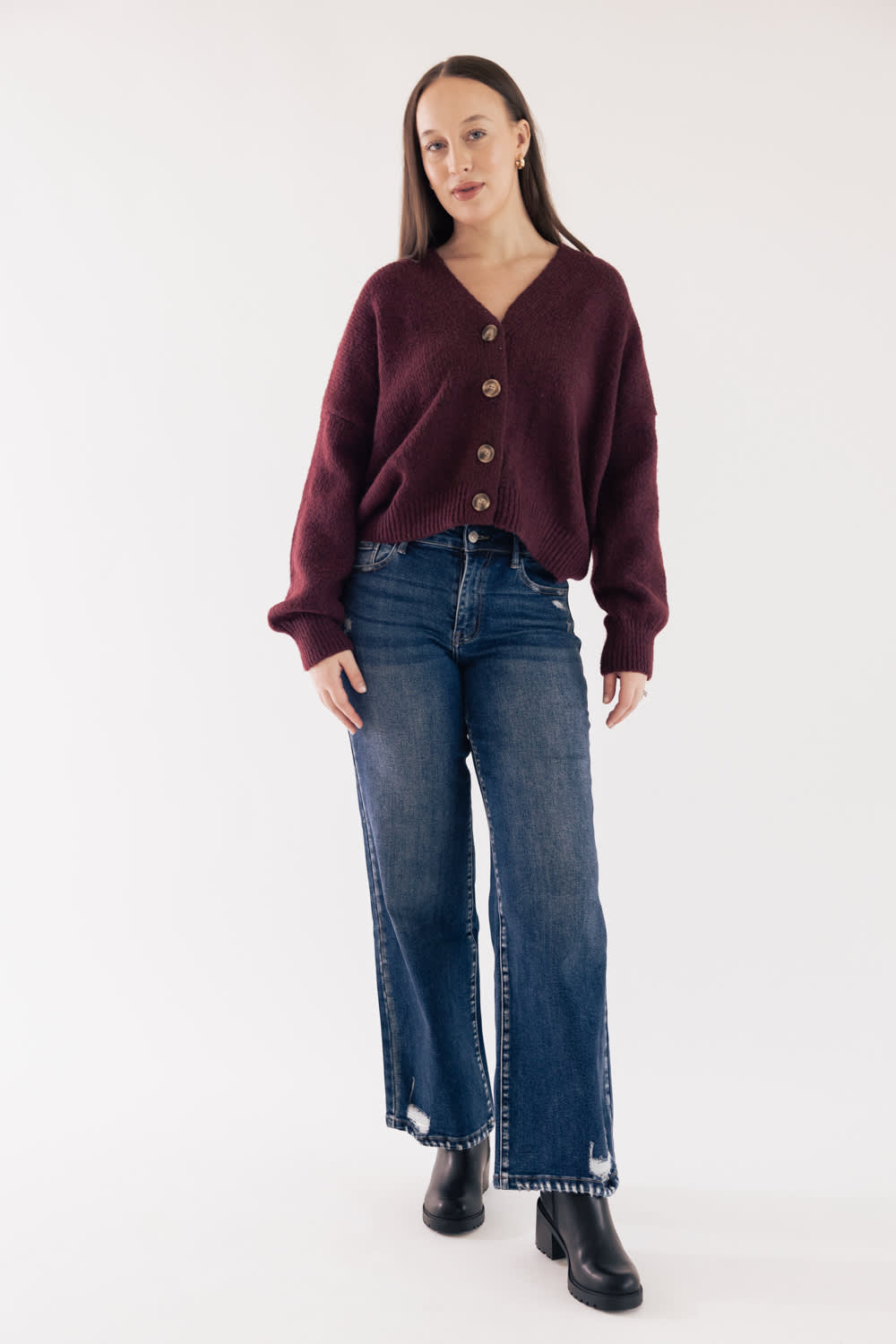 Front view of a model wearing KanCan dark wash, high-rise wide-leg jeans with slight distressing, styled with a button-front burgundy cardigan sweater and black boots against a plain backdrop.