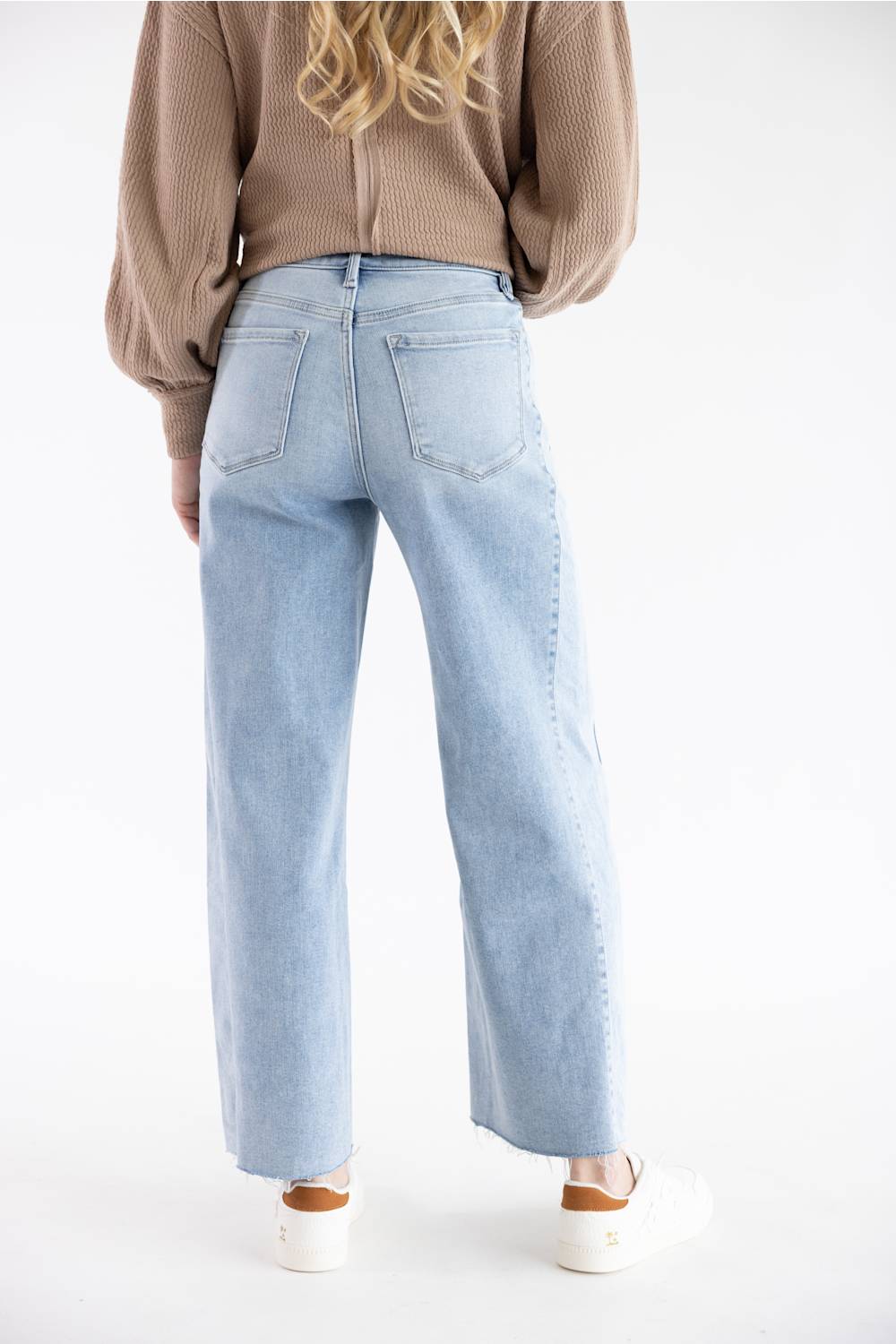 A person shown from the back wearing light-wash wide-leg denim jeans, a brown ribbed sweater, and white sneakers against a plain white studio background.