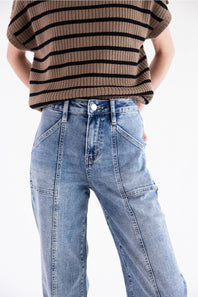 light wash with unique utility-style front pockets and vertical seams down the legs. The model pairs them with a brown and black striped knit top against a clean white studio background.