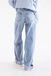 A person shown from the back wearing light-wash blue wide-leg jeans with rolled cuffs, a blue-and-white striped shirt, and white sneakers against a plain white studio background.