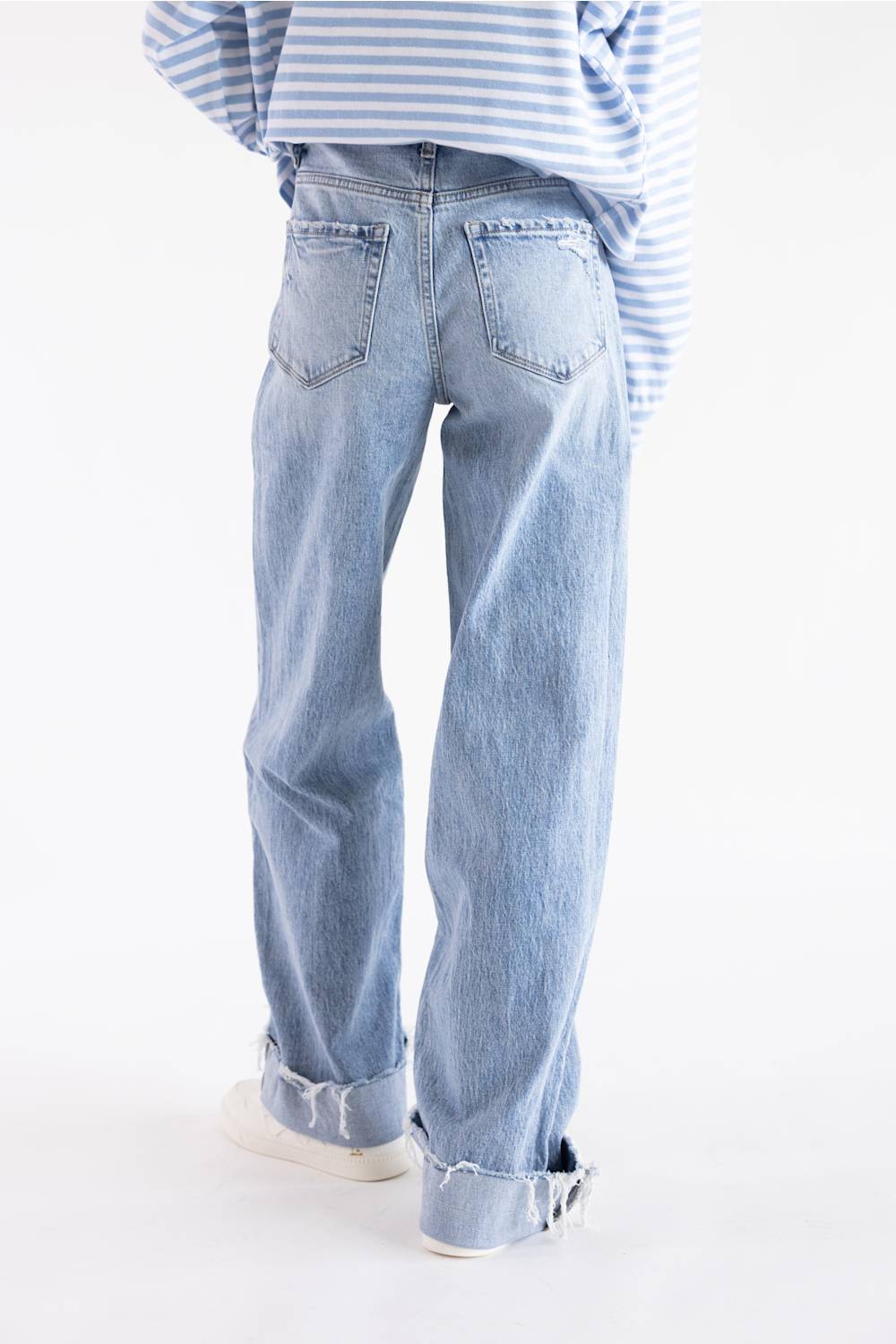A person shown from the back wearing light-wash blue wide-leg jeans with rolled cuffs, a blue-and-white striped shirt, and white sneakers against a plain white studio background.