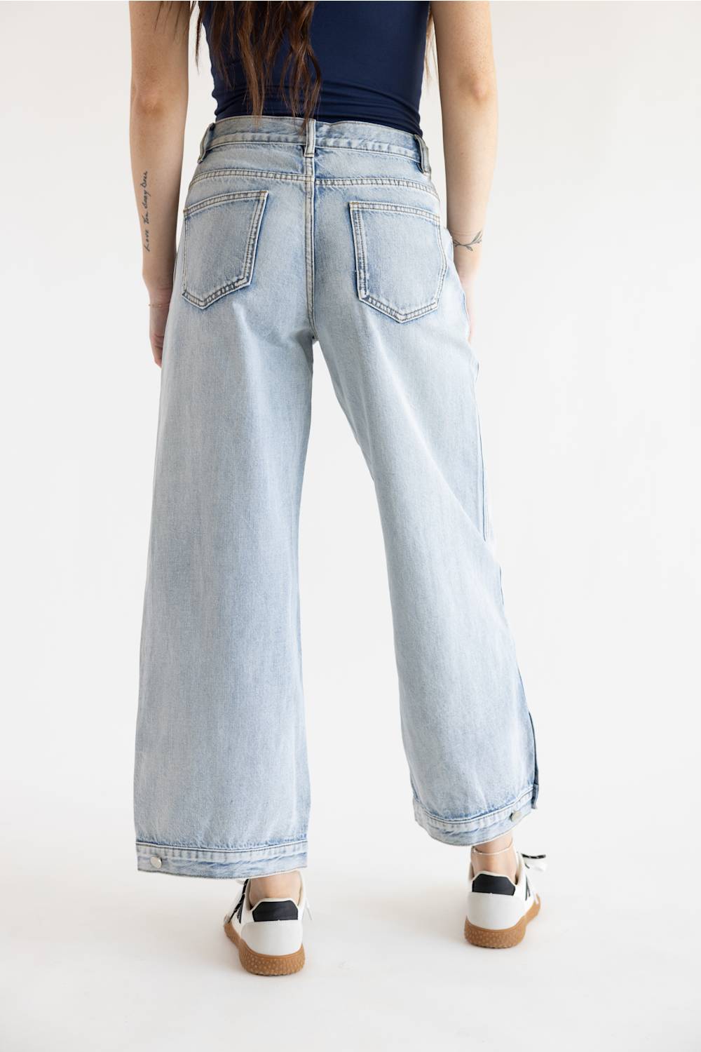 A model posed from the waist down wearing light-wash wide-leg jeans with prominent vertical side seam stitching details, styled with a navy blue fitted top and white sneakers with brown/black accents on a solid white background.
