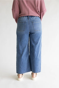 A person shown from the back wearing wide-leg blue denim pants with back patch pockets, a dusty pink top, and white shoes against a plain light background.