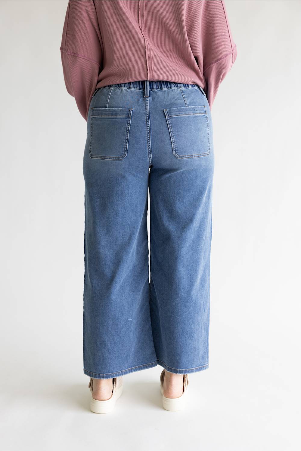 A person shown from the back wearing wide-leg blue denim pants with back patch pockets, a dusty pink top, and white shoes against a plain light background.
