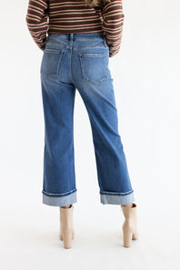 A person shown from the back wearing blue wide-leg cropped jeans with rolled cuffs, a brown striped long-sleeve top, and beige ankle boots against a plain white studio background.