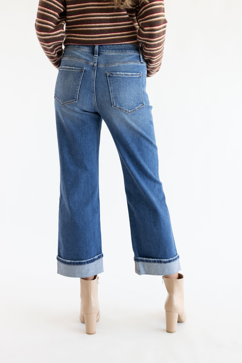 A person shown from the back wearing blue wide-leg cropped jeans with rolled cuffs, a brown striped long-sleeve top, and beige ankle boots against a plain white studio background.