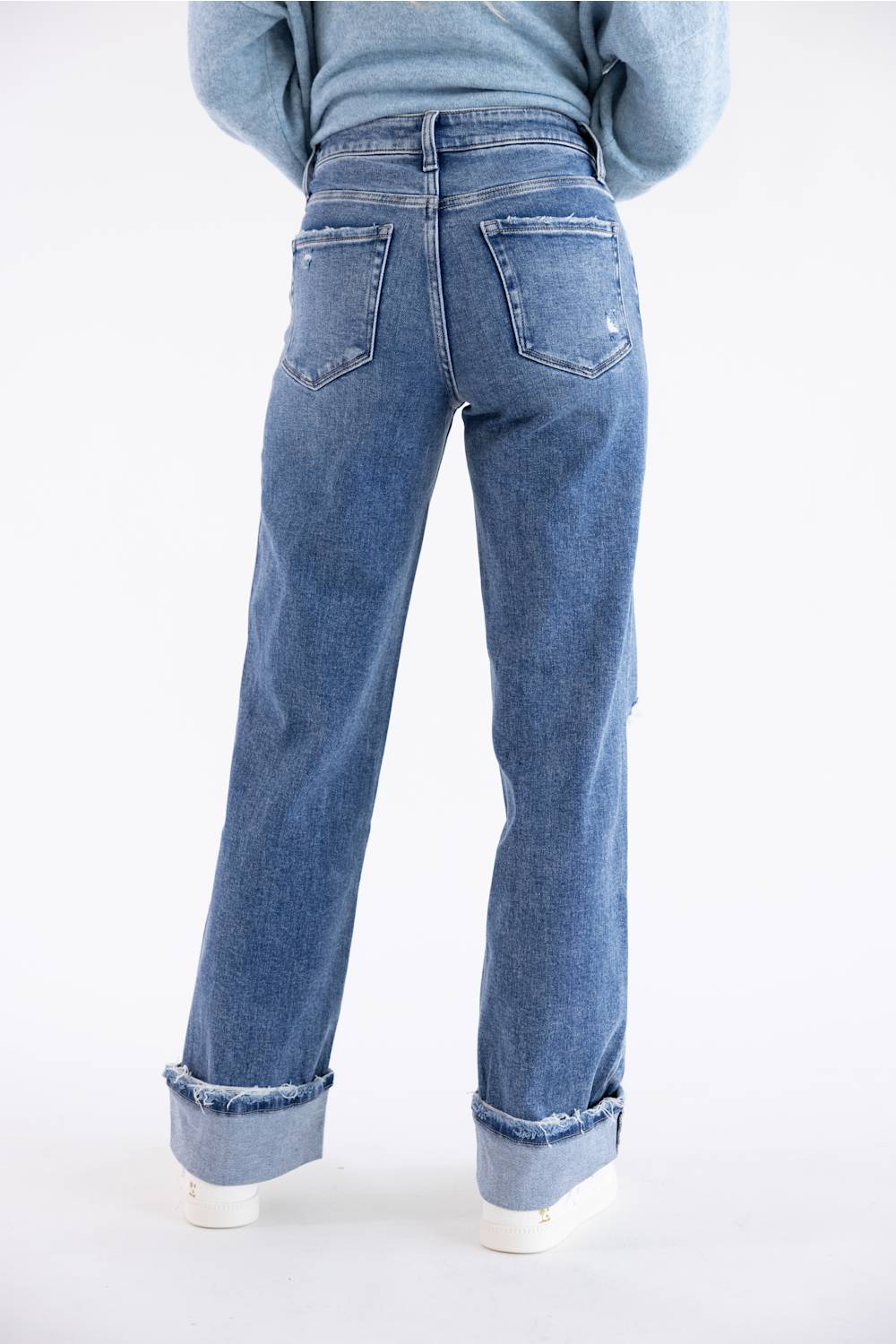 A person shown from the back wearing blue denim jeans with a straight-leg or relaxed fit, cuffed hems, and a light blue sweater against a plain white studio background.