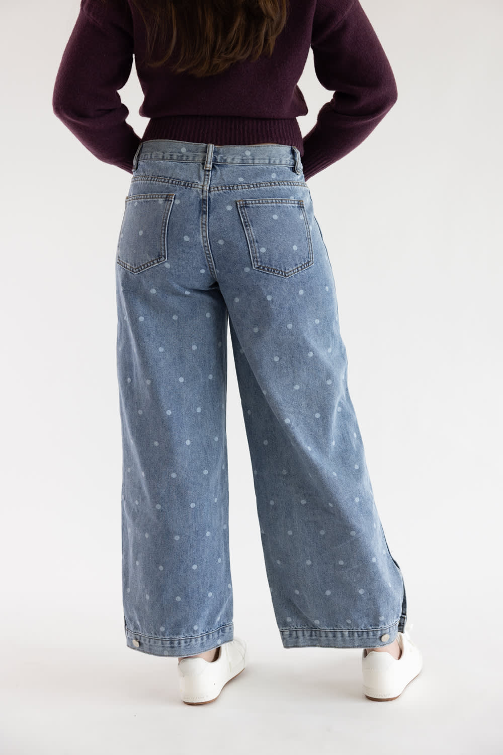 A person shown from the back wearing light-wash blue wide-leg jeans with a small white star print, a dark plum sweater, and white sneakers against a plain white studio background.