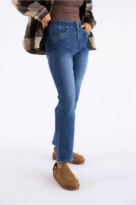 A person wearing medium-wash blue jeans with a brown top, a brown-and-black plaid flannel shirt, and tan slip-on boots, shown from the waist down against a plain white studio background.