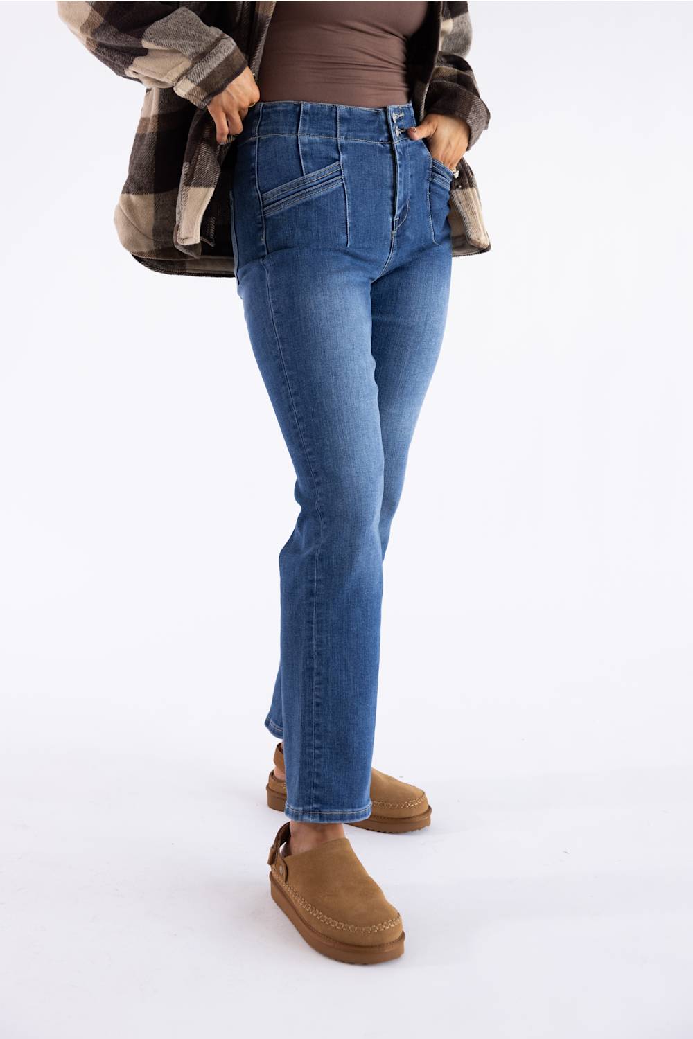 A person wearing medium-wash blue jeans with a brown top, a brown-and-black plaid flannel shirt, and tan slip-on boots, shown from the waist down against a plain white studio background.