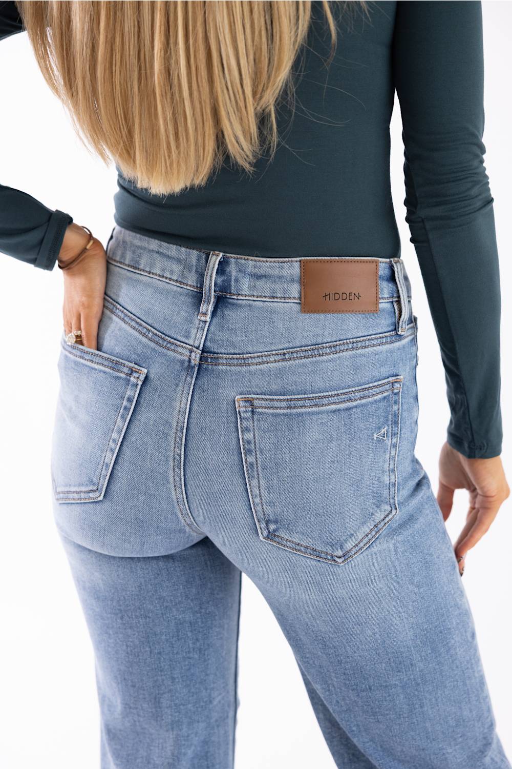 Rear view of a woman wearing light-wash blue jeans with a brown leather brand patch on the back waistband, paired with a dark teal long-sleeve top against a white background.