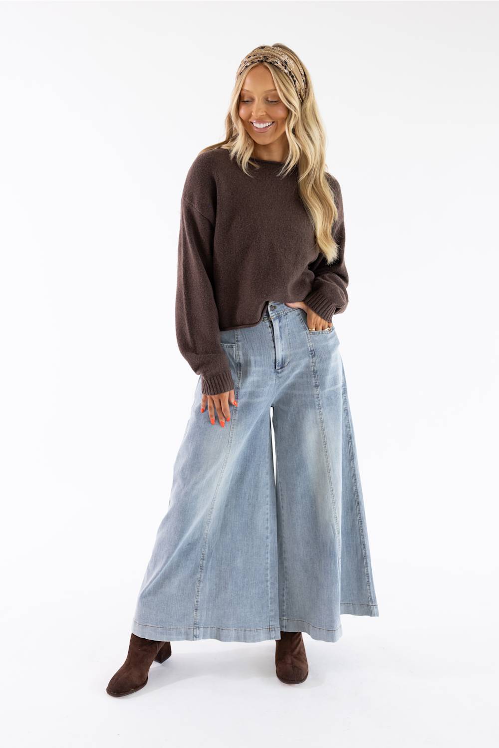 A woman modeling a dark brown crewneck knit sweater and wide-leg light blue denim pants, paired with brown suede boots, against a solid white studio background.