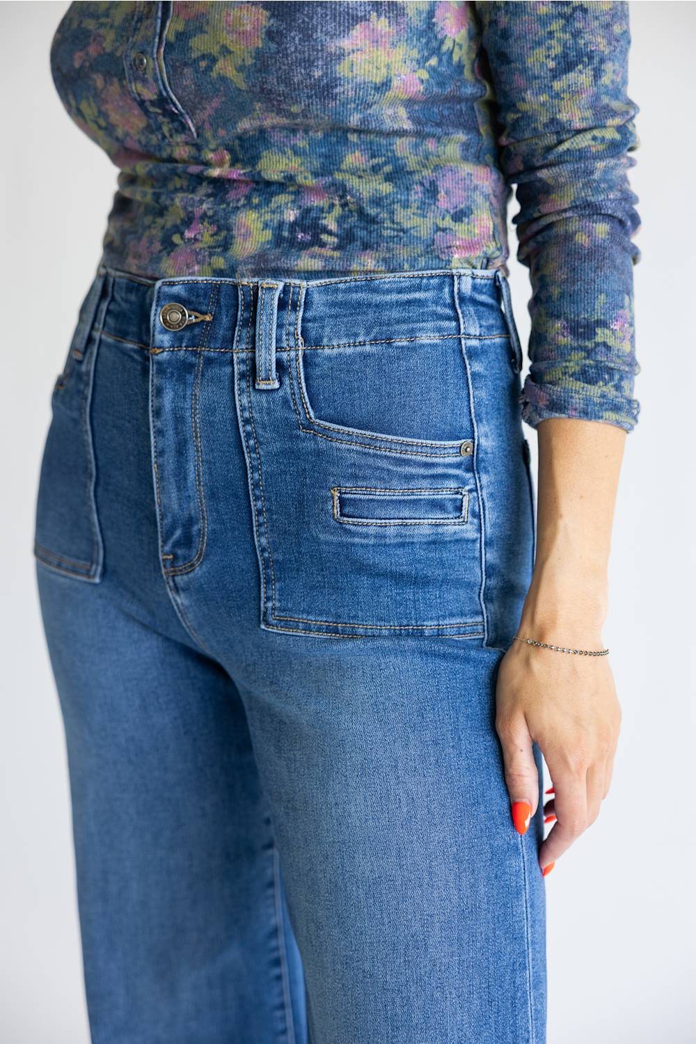 A close-up of a person wearing high-waisted blue denim jeans with a floral long-sleeve top, shown from the chest down against a plain light background.
