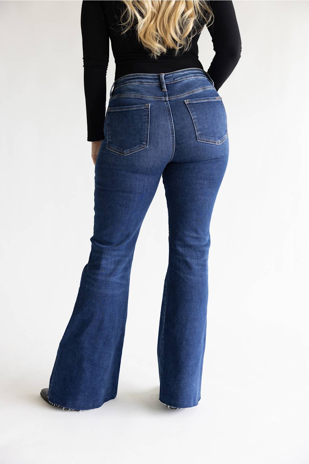 A person shown from the back wearing dark blue flared denim jeans and a black long-sleeve top against a plain light gray studio background.