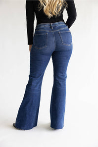 A person shown from the back wearing dark blue flared denim jeans and a black long-sleeve top against a plain light gray studio background.