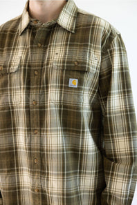 A close-up of a person wearing a Carhartt plaid button-up shirt in brown, beige, and green tones, showing the chest pocket with the signature Carhartt logo patch.