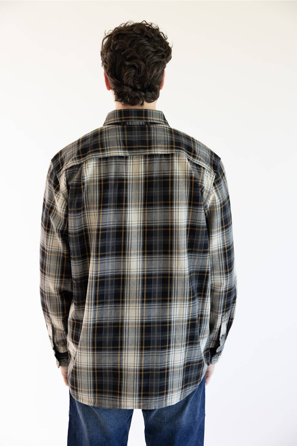 A man viewed from behind wearing a long-sleeve plaid button-up shirt in black, white, and tan, paired with blue jeans against a plain white background.