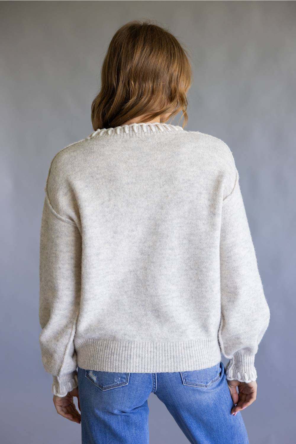 A woman viewed from behind wearing a light heather gray knit sweater and blue jeans, standing against a neutral gray studio background.