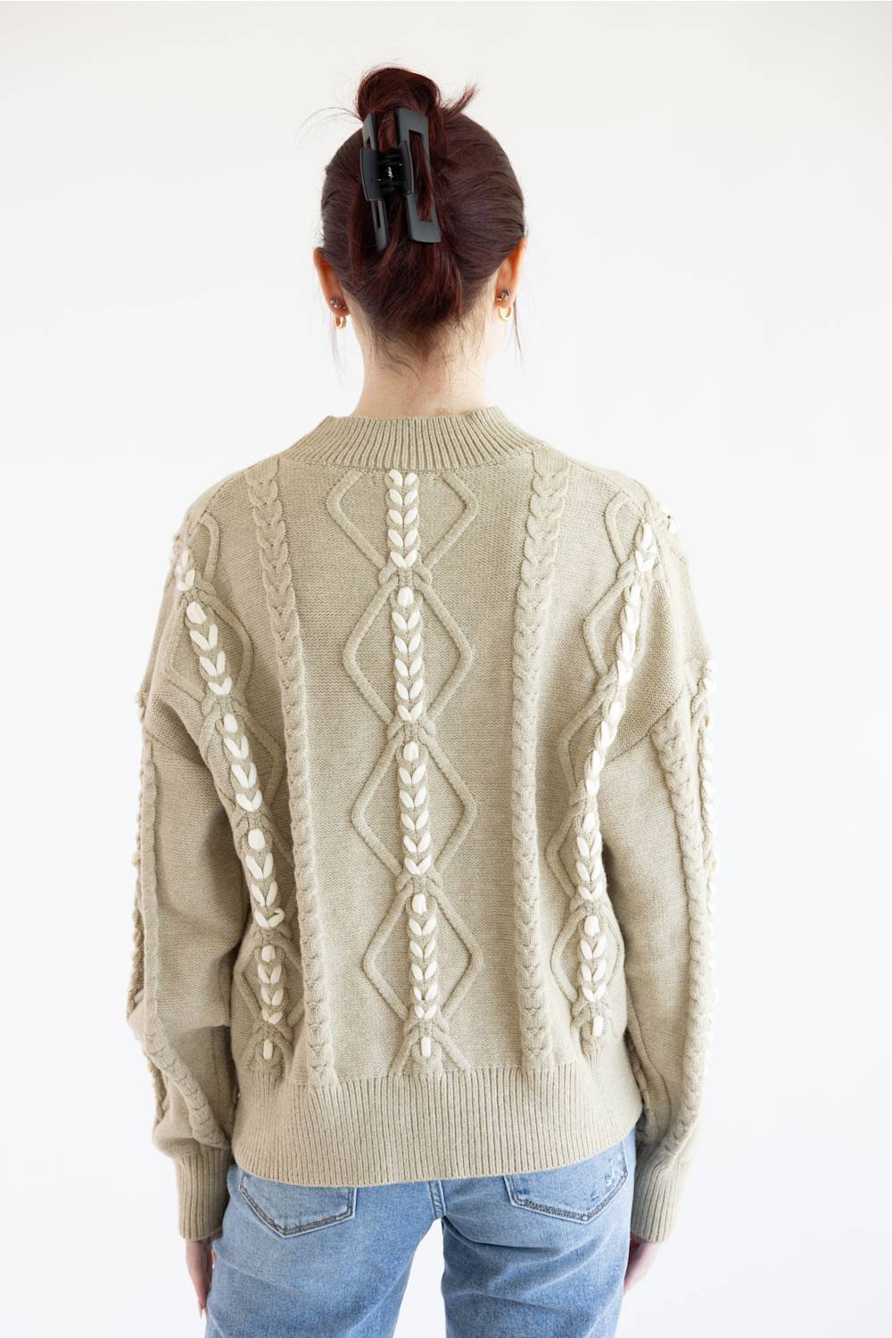 A person shown from the back wearing a cream-colored cable-knit sweater with diamond and braid patterns, paired with blue jeans against a plain white background.