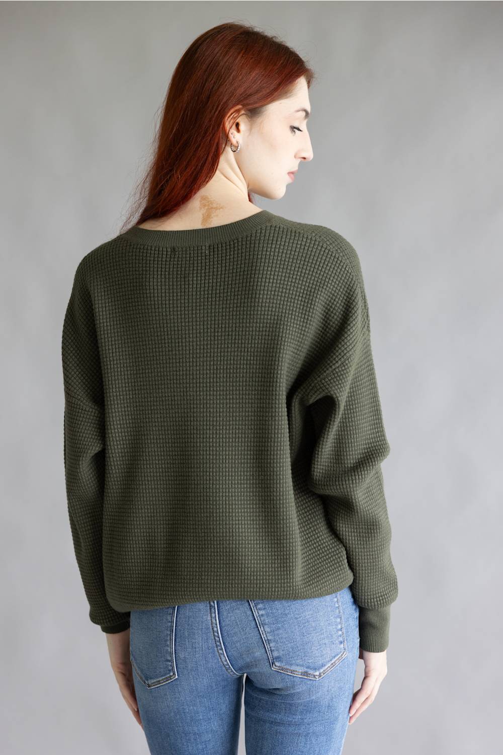 A woman with red hair is shown from the back wearing an olive green waffle-knit sweater and blue jeans against a plain gray studio background.
