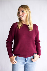 A woman with blonde hair smiling and looking to the side, wearing a textured deep burgundy long-sleeve knit sweater and light blue jeans, standing against a light gray studio background.